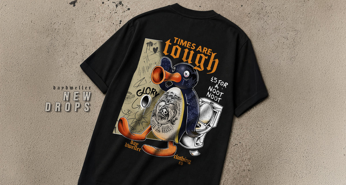 A black short-sleeve graphic T-shirt laid flat on a textured beige background, showing the back design with illustrated penguins, orange text reading 'TIMES ARE TOUGH', and a tattoo-style circular motif. The shirt also has a visible 'baptizedier NEW DROPS' text overlay on the left side.