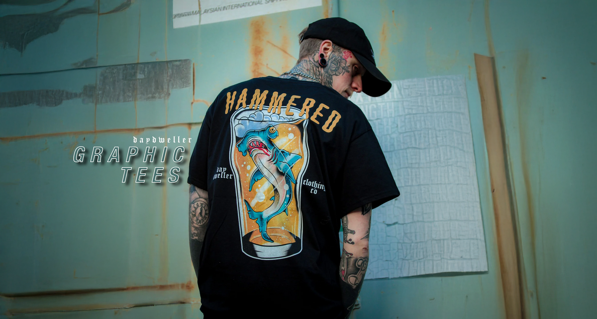 A tattooed man wearing a black graphic t-shirt with a large hammer-themed print on the back, standing against a weathered teal wall.